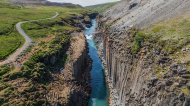Resimde İzlanda 'daki Studlagil Kanyonu, dik bazalt sütunları, turkuaz bir nehir, yeşil tepeler ve manzaradaki dolambaçlı toprak bir yol yer alıyor..