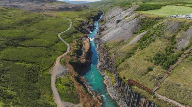 İzlanda 'daki Studlagil Kanyonu' nun dramatik hava manzarası, bazalt sütunlar, turkuaz bir nehir, yeşil bitki örtüsü ve ahşap bir izleme platformu yer alıyor..
