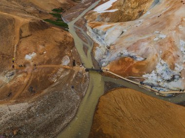 İzlanda 'da turuncu ve beyaz arazi, dolambaçlı nehir, buhar bacaları, kar bantları ve keşif yapan ziyaretçilerin yer aldığı canlı bir jeotermal alanın havadan görüntüsü..
