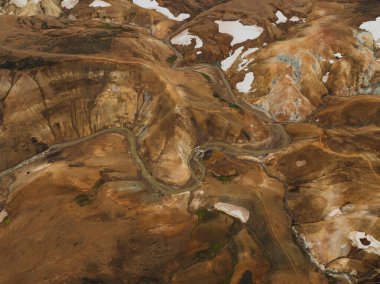 Turuncu, kahverengi ve sarı renkleri olan engebeli rhyolite tepeleri, kar parçacıkları ve dolambaçlı bir dere bu jeotermal İzlanda manzarasını tanımlar..