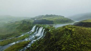 Bir çağlayan şelale, yosun kaplı arazinin ortasındaki dolambaçlı bir nehre akar. Uçurumlar ve sisli tepeler tarafından çerçevelenmiş, aracı olan dar bir yol görülebilir..