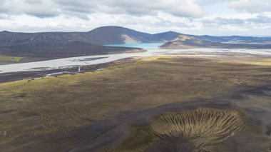 İzlanda volkanik kraterinin, dolambaçlı bir nehrin ve bulutlu bir gökyüzünün altındaki karanlık dağlarla çevrili bir turkuaz gölün hava görüntüsü..