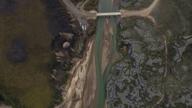 İzlanda 'da yeşil bir nehri geçen küçük bir köprünün hava görüntüsü, bataklık arazi, kumlu alanlar, kayalık zemin ve görünür park edilmiş araçlarla çevrili..