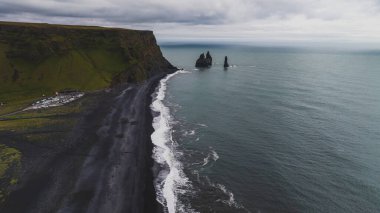 Reynisfjara plajı siyah kum, Reynisdrangar bazalt deniz yığınları, engebeli yeşil uçurumlar, park edilmiş araçlar ve okyanus üzerinde bulutlu bir gökyüzü barındırır..