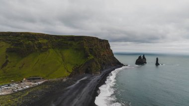 Reynisfjara plajı siyah kum, Reynisdrangar bazalt deniz yığınları, yosun kaplı bir uçurum ve bulutlu gökyüzünün altında görünür araçları olan bir park alanına sahiptir..