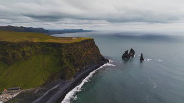 Reynisfjara 'nın dramatik uçurumları ve Reynisdrangar bazalt deniz yığınları, yemyeşil kayalıklar ve kayalıkların tepesinde küçük bir yapı..