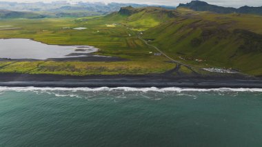 İzlanda 'nın kara kumsalının dramatik hava manzarası, yeşil tepeler, virajlı yollar, dağınık binalar, okyanus dalgaları ve kırsal kesimde küçük bir göl..