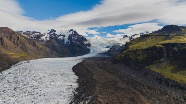 Vatnajokull Ulusal Parkı 'ndaki bir buzulun dramatik hava manzarası, yosun kaplı dağlar ve bulutlu bir gökyüzünün altındaki volkanik arazi arasında buzlar akıyor..