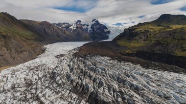 Karanlık çökelti çizgileri olan, yosun kaplı dağlarla çevrili İzlanda buzulunun dramatik hava manzarası ve bulutlu bir gökyüzünün altında kar tepesi..