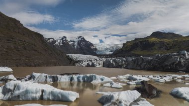 Buzullar, volkanik kayalar, yeşil yosunlar ve parçalı bulutlu gökyüzünün altındaki kar tepeleriyle çevrili, kahverengimsi bir göle doğru uzanır..