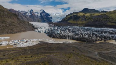 Dağlardan, volkanik arazi, yeşil tepeler ve bulutlu bir gökyüzünün altındaki kar tepeleriyle çevrili buzdağlarıyla dolu çamurlu bir lagüne uzanan bir buzuldur..
