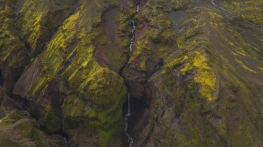 İzlanda 'da yosun kaplı bir kanyonun dramatik hava manzarası, engebeli kaya oluşumlarının ortasındaki karanlık bir havuza dökülen ince bir şelalenin görüntüsü..