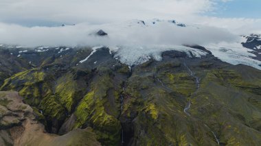İzlanda 'da bir buzul, dar şelaleleri olan yosun kaplı dağlara iner. Alçak bulutlar kısmen buzlu ve volkanik araziyi kaplar..