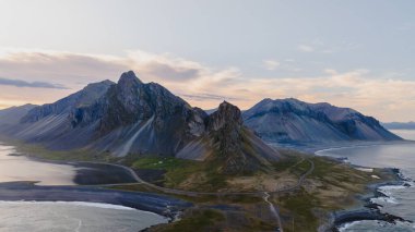 Vestrahorn 'un sarmal tepeleri, kıvrımlı bir yolu, siyah kumlu plajları, İzlanda' da bir lagünü ve Atlantik Okyanusu olan pastel bir gökyüzüne karşı yükselir..