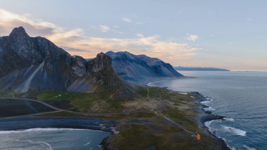 İzlanda 'daki Vestrahorn dağlarının dramatik hava manzarası sivri tepeleri, kıyı yolu, siyah kumlu plajları ve okyanus kıyısında turuncu bir deniz feneri..