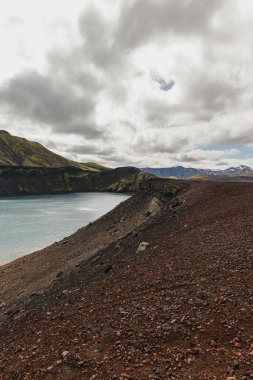Kırmızımsı volkanik toprak, yosun kaplı tepeler ve bulutlu gökyüzünün altındaki karlı dağlarla çevrili sakin bir göl..