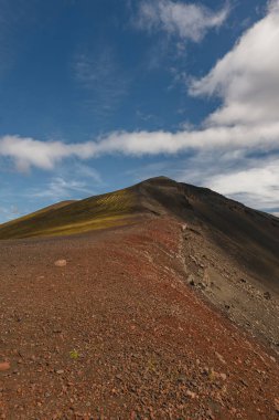 İzlanda 'da volkanik bir tepe, kırmızı ve siyah kayalık arazi, sarı yeşil yosun parçaları, ve dağınık mavi bulutların altında bir tepe..