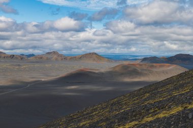 İzlanda 'da küllerle kaplı kıraç bir volkanik arazi, kırmızı renkli tepeler, yosunlu yosunlar, engebeli dağlar ve parçalı bulutlu bir gökyüzünün altında toprak bir yol..