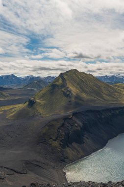 İzlanda 'nın karanlık volkanik arazisinden yükselen yeşil kaplı bir dağ, tabanında parıldayan bir göl, engebeli uçurumlar ve arka planda karlı zirveler var..