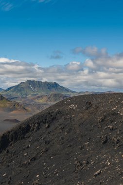 İzlanda 'nın engebeli volkanik arazisi. Arkasında yeşil bir dağ var. Parlak mavi bir gökyüzünün altında karla kaplı..