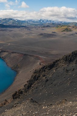 Canlı bir mavi krater gölü kayalık volkanik arazi, yosun kaplı tepeler ve parçalı bulutlu bir gökyüzünün altında karla kaplı dağlar ile çevrilidir..