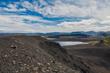 Volkanik yamaçlar, yosun ve çorak topraklarla çevrili, parçalı bulutlu bir gökyüzünün altında uzak dağlarla çevrili yansıtıcı bir göle uzanır..