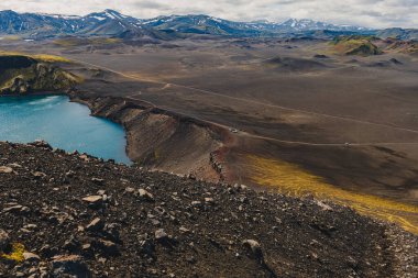 Karanlık volkanik zemin, sarı yosun ve İzlanda 'da parçalı bulutlu bir gökyüzünün altındaki uzak kar kaplı dağlarla çevrili canlı bir mavi krater gölü..