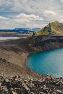 Turkuaz krater gölü siyah volkanik kayalar ve yeşil yosun kaplı dağlarla çevrili, karla kaplı zirveler ve ıssız arazi.