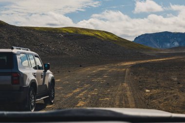 İzlanda 'nın volkanik dağlık arazilerinde engebeli bir toprak yolda gümüş renkli bir SUV park halinde. Sahnede kayalık arazi, yosun kaplı tepe ve uzak dağlar yer alıyor..