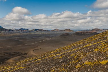 İzlanda 'da verimsiz bir volkanik arazi. Koyu kayalar, sarı yeşil yosunlar, dolambaçlı toprak bir yol ve mavi gökyüzünün altındaki engebeli dağlar..