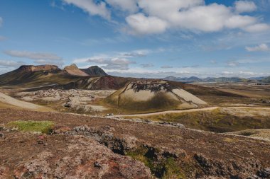 Çok renkli ryolit dağları, dolambaçlı toprak yolu, yosunlu kayalık çıkıntıları ve parçalı bulutlu gökyüzünün altındaki uzak tepeleri olan engebeli İzlanda manzarası..