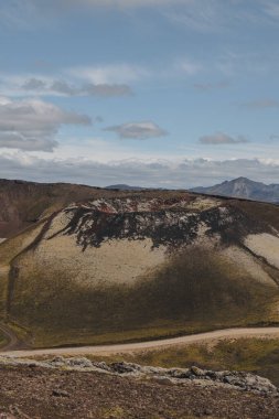 İzlanda 'da, engebeli araziyle çevrili karanlık kaya ve ışık yosunlarıyla çevrili dairesel bir volkanik krater. Arka planda dağlar olan toprak bir yol kıvrılır..