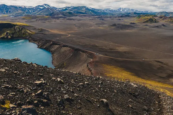 Karanlık volkanik zemin, sarı yosun ve İzlanda 'da parçalı bulutlu bir gökyüzünün altındaki uzak kar kaplı dağlarla çevrili canlı bir mavi krater gölü..