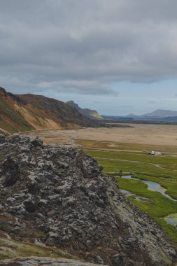 İzlanda 'nın Landmannalaugar bölgesindeki bulutlu bir gökyüzünün altında koyu volkanik kayalar yeşil çayırlar, dolambaçlı dereler ve renkli rhyolite dağları oluşturur..