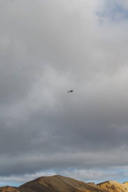 Bir helikopter çorak tepelerin üzerinde bulutlu bir gökyüzüne doğru uçuyor, İzlanda 'nın volkanik manzarasını yeryüzü tonları ve izolasyon hissi ile sergiliyor..