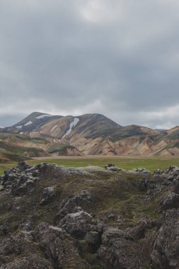 Turuncu, kahverengi ve yeşil renkli ryolit dağları, kar yamaçları ve İzlanda 'da bulutlu bir gökyüzünün altında yosun kaplı lav tarlaları.
