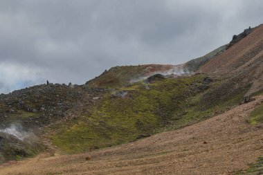 İzlanda 'da buharlı bacaları, yosun kaplı tepeleri, kırmızımsı kahverengi çakılları ve bulutlu bir gökyüzünün altındaki tepede yalnız bir figür olan engebeli bir jeotermal arazi..