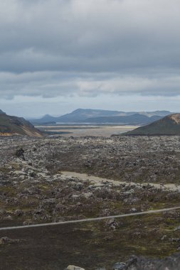 İzlanda 'daki volkanik bir arazide karanlık lav tarlaları, yosun yamaları, geniş bir vadi ve bulutlu bir gökyüzünün altında uzak dağlar bulunur..