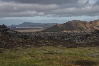 İzlanda 'nın engebeli arazisi yosun kaplı kayalar, pürüzlü lav tarlaları ve bulutlu bir gökyüzünün altında çizgili renkli dağlar..