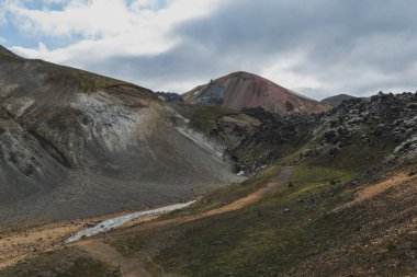 Renkli rhyolite dağları, kırmızımsı bir tepe, dolambaçlı bir dere, ve Landmannalaugar 'da işaretli bir yürüyüş yolu bulutlu bir gökyüzü altında.