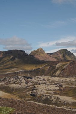Çok renkli ryolit dağları, volkanik kayaları kaplayan yosunlar ve dağınık bulutlu mavi gökyüzünün altında merkezi bir tepe ile engebeli İzlanda manzarası..