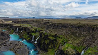 İzlanda 'daki dramatik kanyonda çağlayan şelaleler, turkuaz bir nehir, yosun kaplı uçurumlar ve bulutlu bir gökyüzünün altındaki dağ sıraları yer alıyor..