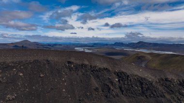 Karanlık kayalık toprak, seyrek bitki örtüsü, dolambaçlı nehirler, uzak dağlar ve parçalı bulutlu gökyüzü ile İzlanda 'nın volkanik arazisinin dramatik hava manzarası..