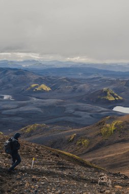 Yalnız bir yürüyüşçü yeşil yosunları, yuvarlanan tepeleri ve muhtemelen İzlanda 'da bulutlu bir gökyüzünün altında küçük bir gölü olan engebeli bir volkanik arazide dolaşır..