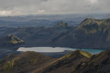 Yeşil yosunlu karanlık volkanik arazi, yansıtıcı bir göl ve bulutlu bir gökyüzünün altında görülebilir dağ zirveleri olan turkuaz bir su kütlesi..