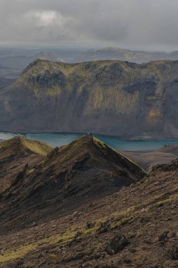 Turkuaz bir nehir, İzlanda 'da yosun kaplı uçurumların altından akar. Mavi ceketli yalnız bir figür bulutlu bir gökyüzünün altındaki bayırda duruyor..