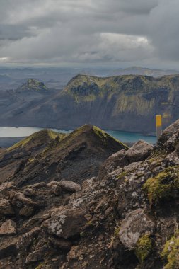 Volkanik bölgede yosun kaplı kayalar, turkuaz bir göl ve bulutlu bir gökyüzünün altında dramatik dağ tepeleri. Sarı işaret direği görünüyor..