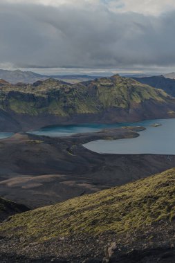 İki turkuaz göl, yosun kaplı tepelerin ve karanlık volkanik toprağın arasında, bulutlu bir gökyüzünün altında, İzlanda 'nın engebeli ve uzak manzarasını gözler önüne seriyor..