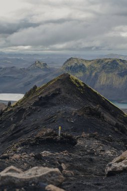 Karanlık bir volkanik tepe yosunlu yosunlu yamaçlar boyunca uzanır, dağlar ve arka planda yansıtıcı bir göl, bulutlu bir gökyüzünün altında. Bir iz işareti görünür.