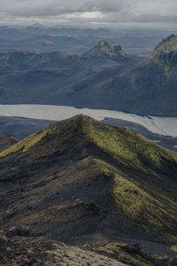 Yeşil yosunlarla kaplı engebeli bir volkanik sırt karanlık toprağa zıt düşer. Soluk bir buzul nehri ise sivri tepelerin ve bulutlu gökyüzünün altındaki bir vadide süzülür..